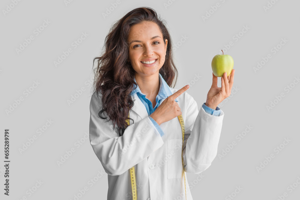 Cheerful young lady doctor in uniform pointing finger on apple isolated on light grey background, smiling at camera