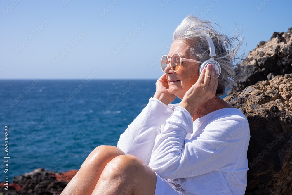 Portrait of smiling senior woman in white listening music with headphones sitting close the beach with closed eyes, elderly lady enjoys retirement or vacation at sea in sunny day