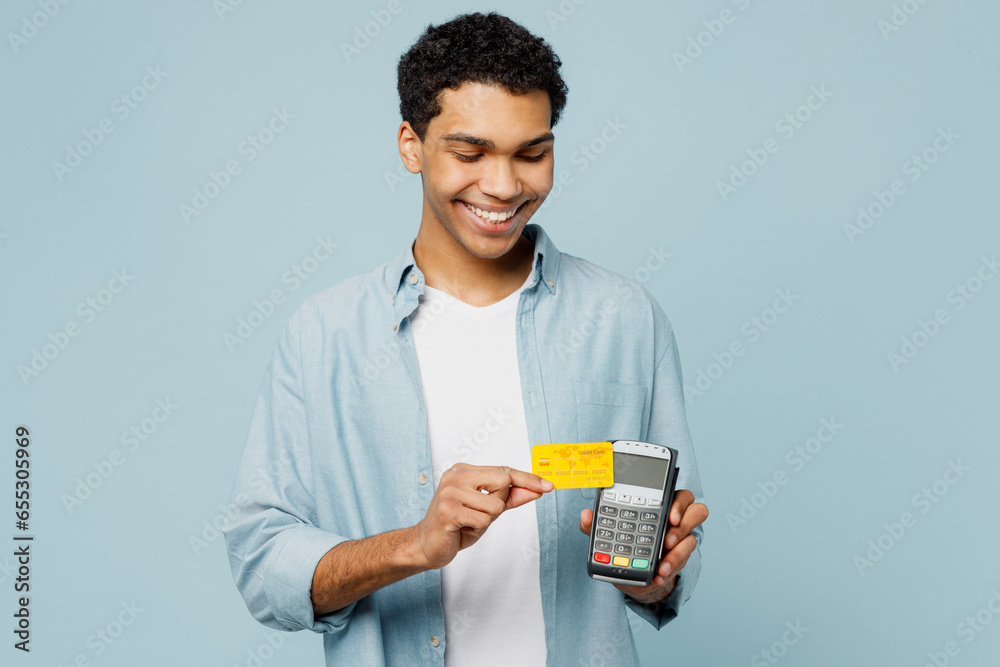 Young man of African American ethnicity wear shirt casual clothes hold wireless modern bank payment terminal to process acquire credit card isolated on plain pastel light blue cyan background studio. Young man of African American ethnicity wear shirt casual clothes hold wireless modern bank payment terminal to process acquire credit card isolated on plain pastel light blue cyan background studio.