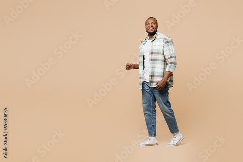 Full body side profile view happy young man of African American ethnicity he wear light shirt casual clothes walking going stroll isolated on plain pastel beige background studio. Lifestyle concept.