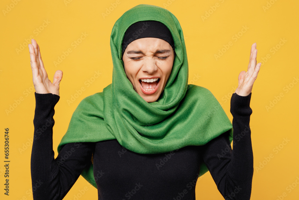 Young angry sad mad arabian asian muslim woman wear green hijab abaya black clothes spread hands scream shout cry isolated on plain yellow background People uae middle eastern islam religious concept