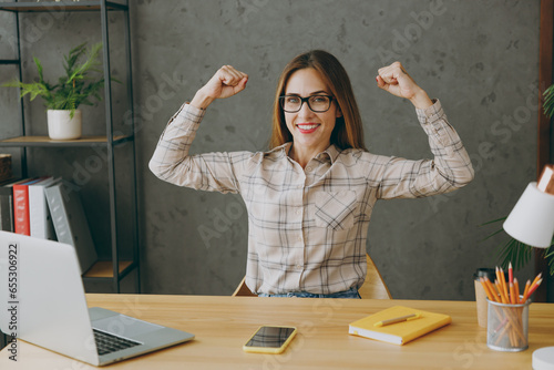 Fototapete Young fitness employee business woman wear shirt casual clothes glasses sit work at office desk with pc laptop show biceps muscles on hand demonstrating strength power
