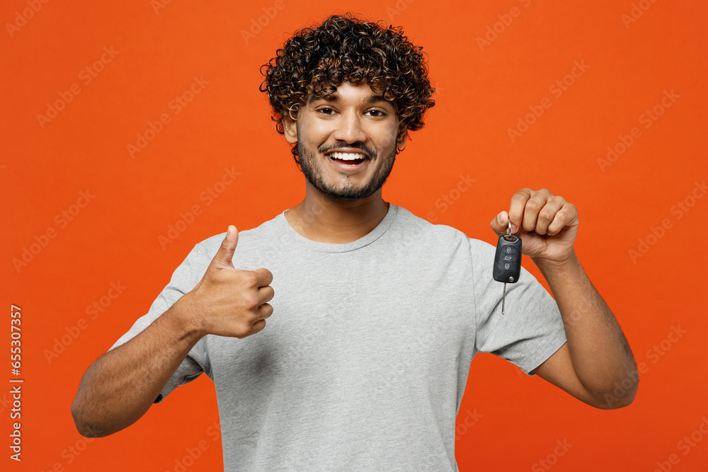 Young smiling happy fun Indian man he wearing t-shirt casual clothes hold in hand car key fob keyless system show thumb up isolated on orange red color background studio portrait. Lifestyle concept. Young smiling happy fun Indian man he wearing t-shirt casual clothes hold in hand car key fob keyless system show thumb up isolated on orange red color background studio portrait. Lifestyle concept.
