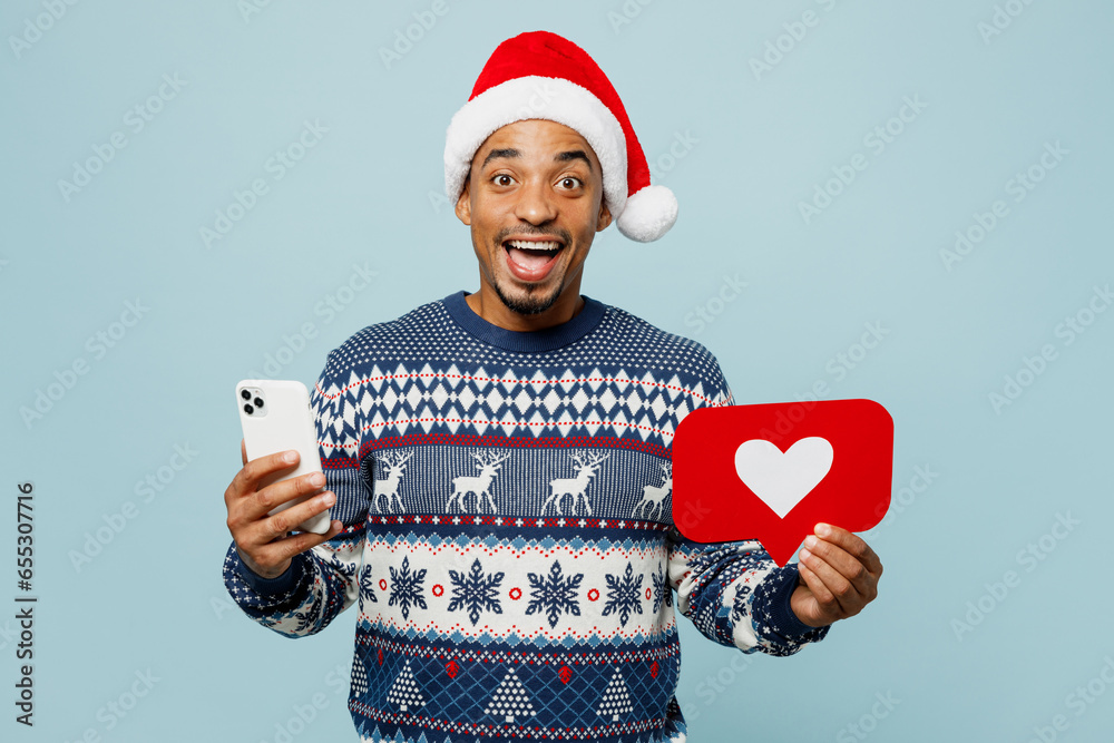 Young man wears knitted sweater Santa hat posing hold mobile cell phone heart form like icon sign isolated on plain pastel blue background. Happy New Year 2024 celebration Christmas holiday concept. Young man wears knitted sweater Santa hat posing hold mobile cell phone heart form like icon sign isolated on plain pastel blue background. Happy New Year 2024 celebration Christmas holiday concept.