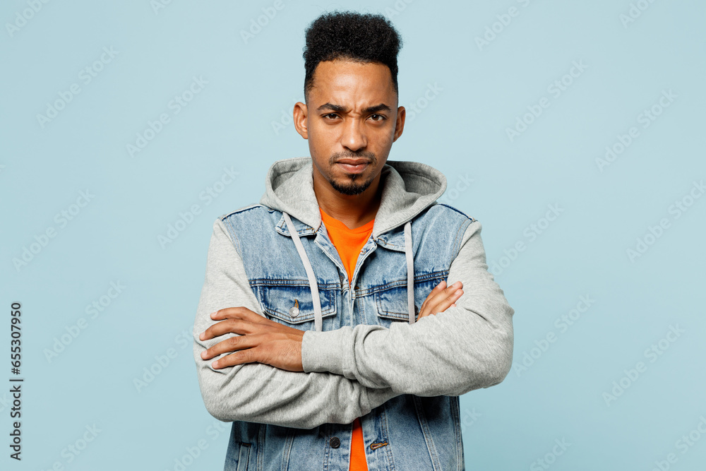 Young sad man of African American ethnicity in denim jacket orange t-shirt hold hands crossed folded look camera isolated on plain pastel light blue cyan background studio portrait Lifestyle concept