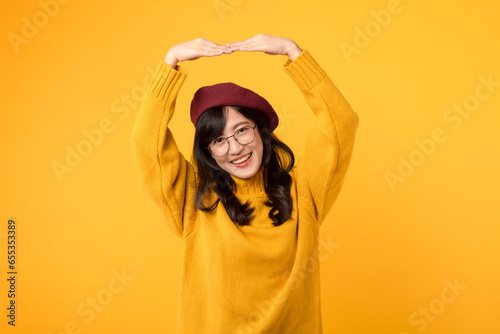 Express your love with a heart arm gesture. A stylish woman 30s in a yellow sweater, red beret, and eyeglasses against a vibrant yellow background. © Jirawatfoto