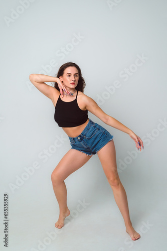 dancehall dancer posing on gray background in the studio, copy space. Full length portrait of beautiful young woman wearing black top and short denim shorts, barefoot
