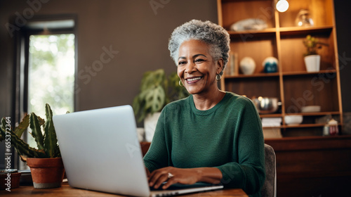 A happy cheerful African-American woman in her 60s with a laptop sits at her desk, smiling, looking at the screen, writing something down, making a shopping list