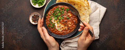 Female hands holding a bowl and eating traditional Indian Punjabi dish Dal makhani with lentils and beans served with naan flat bread, fresh cilantro on brown concrete rustic table top view. photo