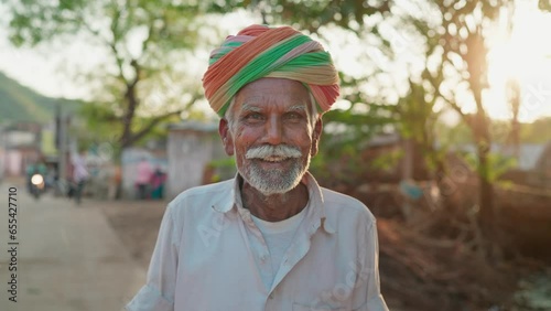 An Indian ethnic happy man or male is wearing a traditional turban or headwear facing or looking at the camera and smiling while standing outdoors in a village