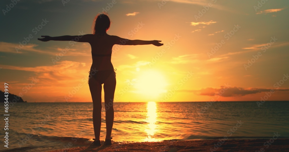 Woman enjoys amazing colorful sunset, standing on a tropical beach. Slim girl silhouette in bikini raises hands against bright orange dramatic sky and ocean waves. Holiday, travel, recreation concept