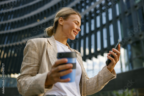 Smiling woman office worker is standing on modern building background and looking on phone photo