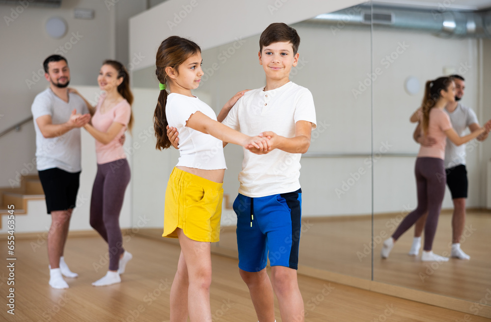 Positive friendly teen sister and brother learning to dance waltz in pair in studio against backdrop of dancing parents. Family bonding and active lifestyle concept..
