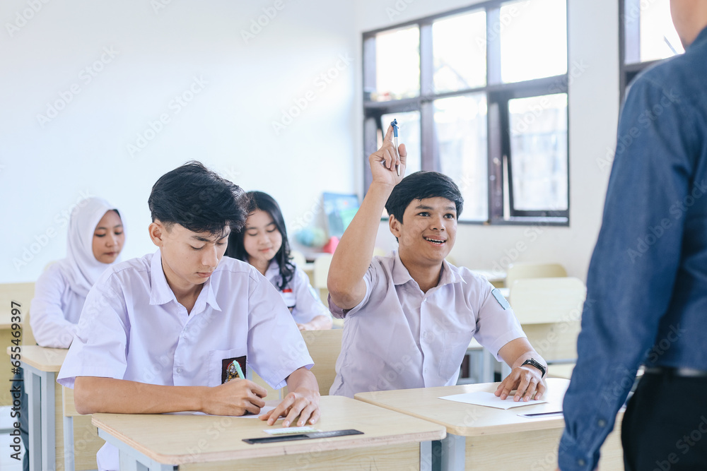 Young Asian high school student in uniform raising hands to answer question from the teacher at the classroom