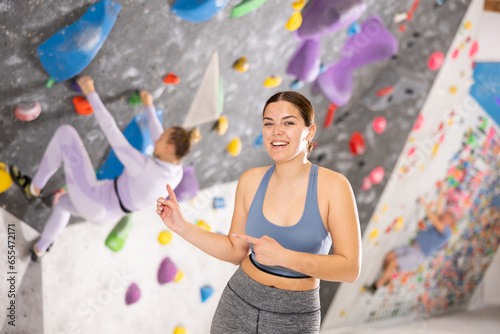 Smiling friendly young girl, climbing coach, showing modern indoor bouldering wall, inviting to training climb