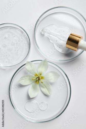Petri dishes with samples of cosmetic oil, pipette and beautiful flower on white background, flat lay