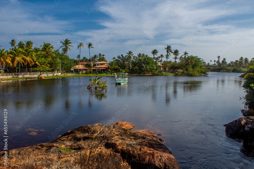 View of the Imbassai River with a boat sailing and reddish rocks, paradisiacal village, on the coast of Bahia, Brazil, near Salvador and Praia do Forte