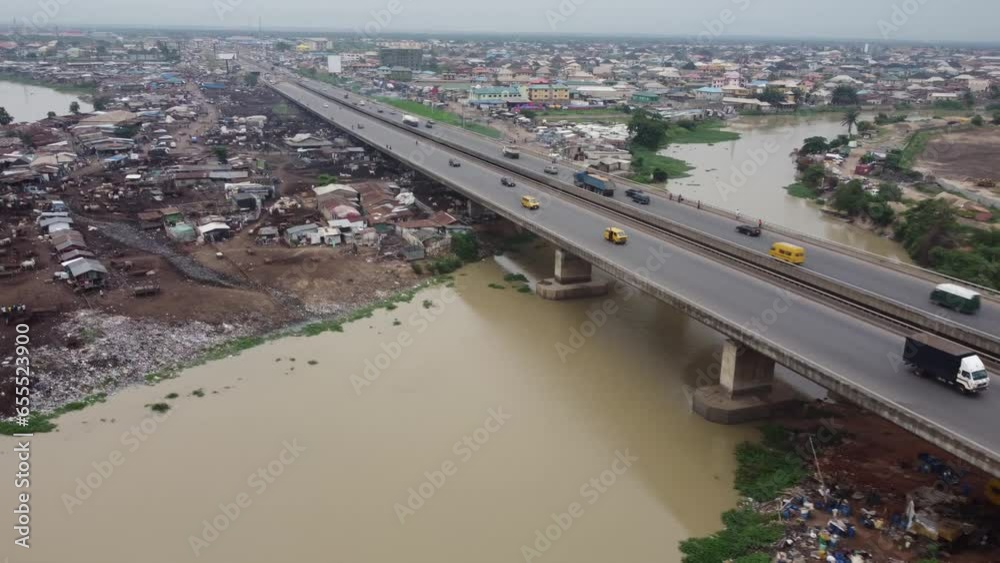Cars moving speedily on brown river bridge of a sub-urban highway with a Nigerian flag in front of an industry