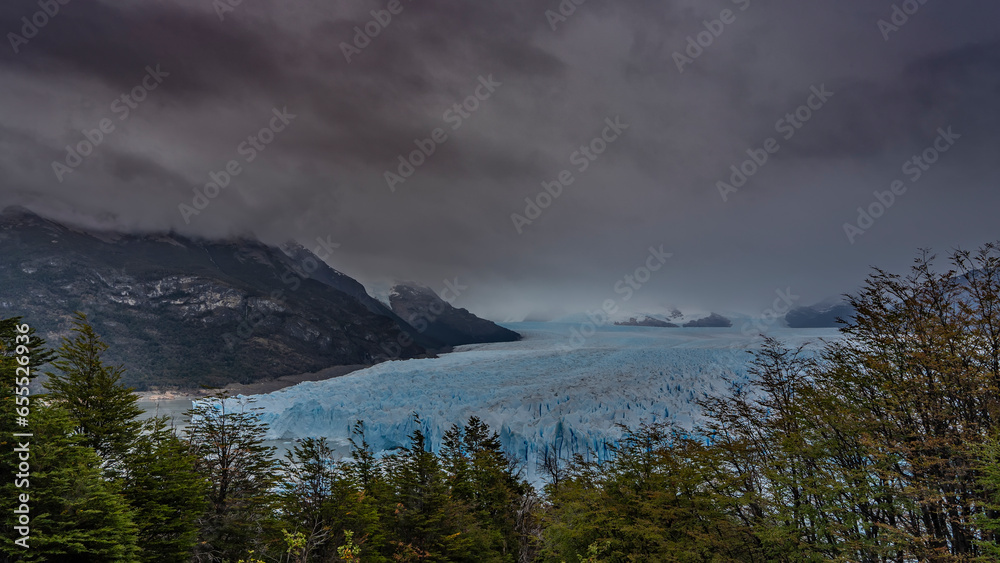 Obraz premium The impressive Perito Moreno Glacier stretches to the horizon between the mountains. A mass of blue ice with sharp peaks, cracks, crevices. Cloudy. Green vegetation in the foreground. El Calafate. 