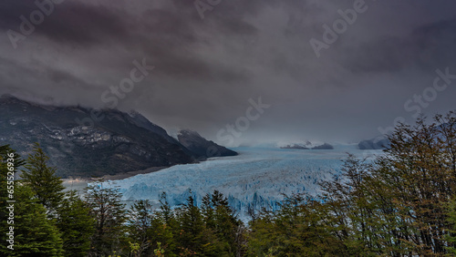 The impressive Perito Moreno Glacier stretches to the horizon between the mountains. A mass of blue ice with sharp peaks, cracks, crevices. Cloudy. Green vegetation in the foreground. El Calafate. 