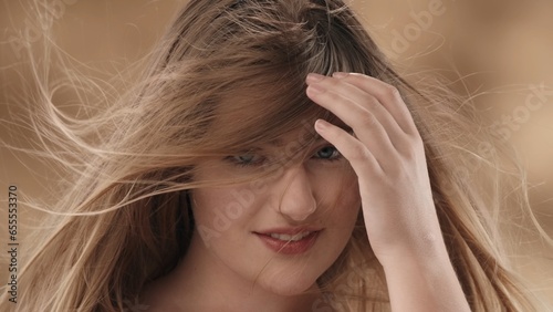 There is a close-up of a young, blonde-haired woman. She stares intently at the camera and smiles. The wind is blowing her hair around her. They cover her face, she removes them with her hand