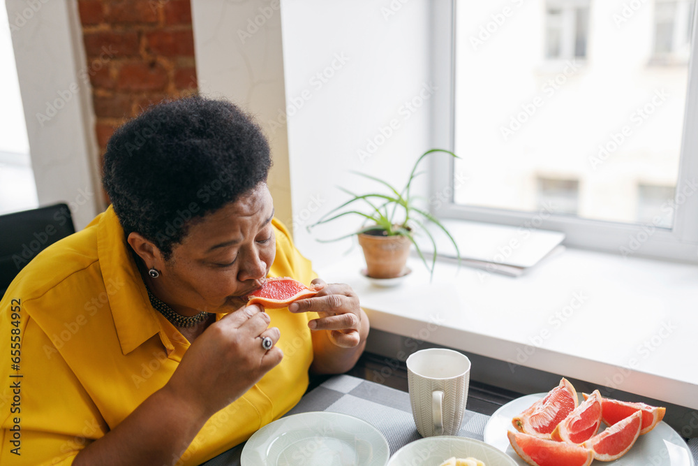 Upper side view image of plus size african american senior retired woman eating juicy fresh organic grapefruit sitting at table near window, being on diet, loosing weight to be healthy
