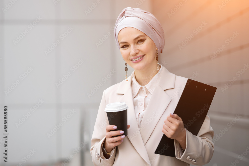 Muslim businesswoman walks past city buildings with black clipboard and carton cup. Woman in silver hijab drinks beverage from paper mug, sunlight Muslim businesswoman walks past city buildings with black clipboard and carton cup. Woman in silver hijab drinks beverage from paper mug, sunlight