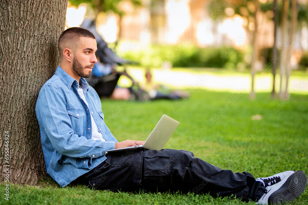 Full length of caucasia young man sitting in a public park and using laptop for e-learning Full length of caucasia young man sitting in a public park and using laptop for e-learning