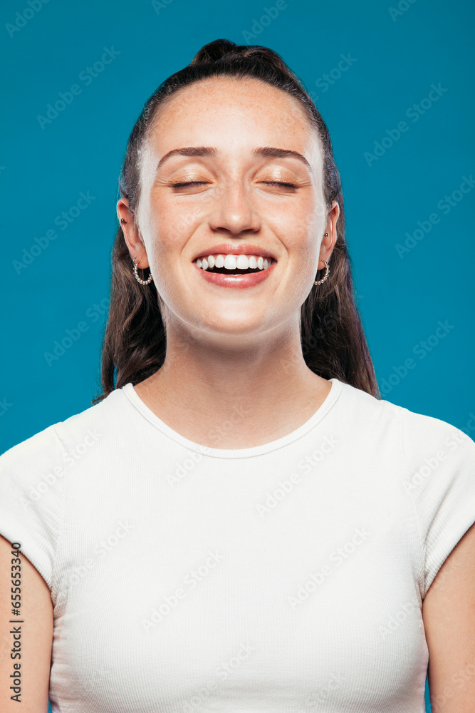 Cheerful female teenager model in casual summer clothes standing in blue studio with eyes closed. Young woman broadly smiles. Cheerful female teenager model in casual summer clothes standing in blue studio with eyes closed. Young woman broadly smiles.