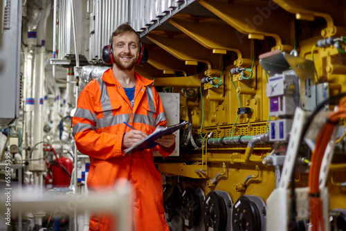 Portrait of young engineer with smile in orange coverall  doing check list in engine room. photo