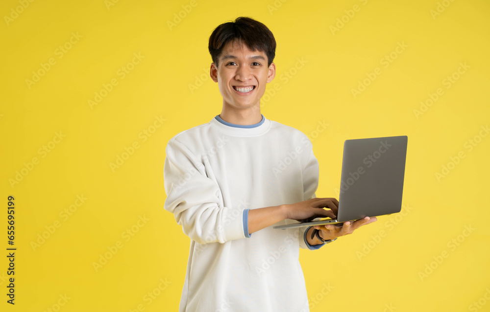 portrait of young Asian man wearing sweater and using laptop on yellow background portrait of young Asian man wearing sweater and using laptop on yellow background