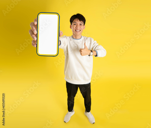 full body of young Asian man wearing sweater and using phone on yellow background