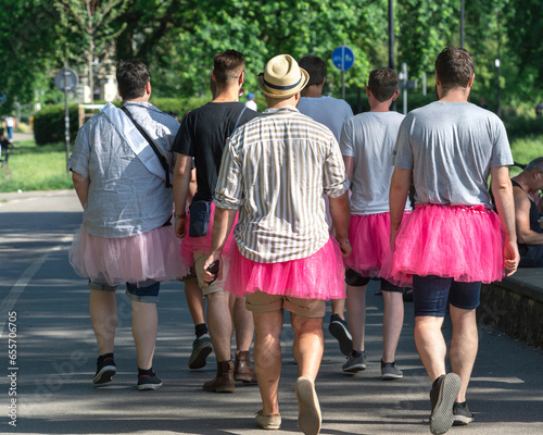 Fototapeta Naklejka Na Ścianę i Meble -  Playful men in vibrant pink tutus stroll in the park, celebrating diversity and pride in the LGBT community. Colorful, whimsical, and fun. Sunny day, view from the back