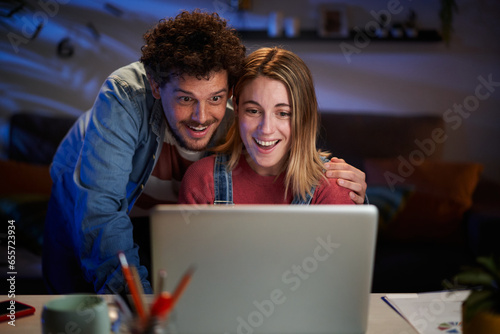 Surprised Caucasian couple looking happy at computer sitting at desk living room at night. Excited millennial people having fun at home using laptop. Positive relationships and leisure moments.