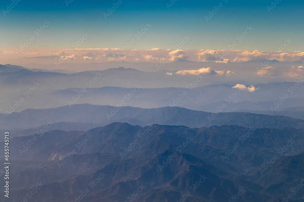 Aerial view of dusky layers of blue and green mountain ranges amid clear blue sky captured from Airplane window