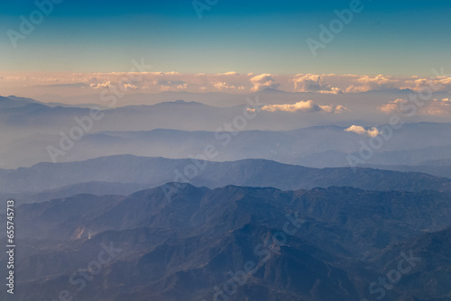 Aerial view of dusky layers of blue and green mountain ranges amid clear blue sky captured from Airplane window