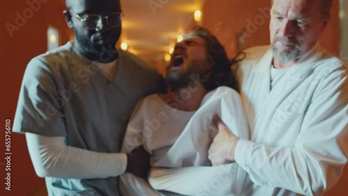 Slow motion shot of two doctors working in asylum restraining violent patient in straitjacket, taking him away along corridor