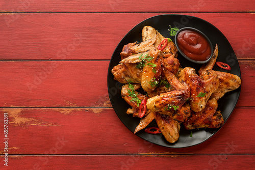 Chicken wings. Grilled or baked chicken wings with sesame seeds and ketchup or spicy tomato sauce on black plate on old wooden red table background. Top view with copy space. © kasia2003