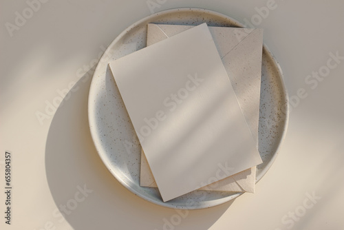 Summer minimal wedding stationery. Blank vertical greeting card mockup with beige envelope, invitation. Ceramic dessert plate in golden sunlight. Table background with shadows overlay. Flatlay, top