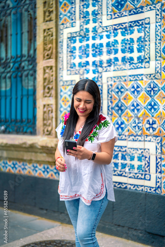 Happy young mexican woman in embroidered top looking at screen of smartphone while standing in front of Sanborns restaurant in downtown Mexico City and reading text messages in daylight photo