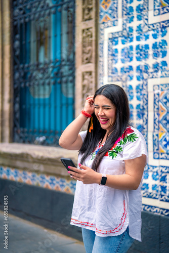 Happy young mexican woman in embroidered top looking at screen of smartphone while standing in front of Sanborns restaurant in downtown Mexico City and reading text messages in daylight photo