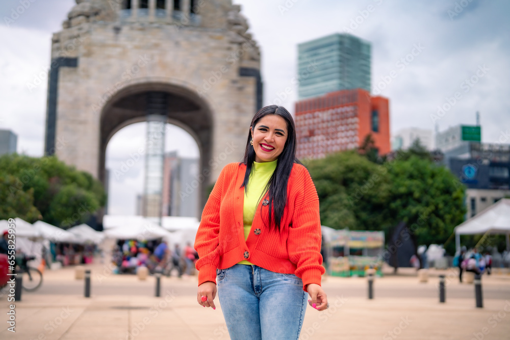 Smiling young Hispanic ethnic woman tourist smiling and looking at camera while standing in street in front of blurred Monument to the Revolution in Mexico city against cloudy blue sky in daylight