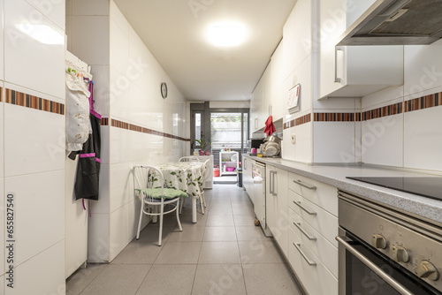 Fototapeta Naklejka Na Ścianę i Meble -  Elongated kitchen with a wall full of white furniture on one side with gray countertop and dining table with matching chairs