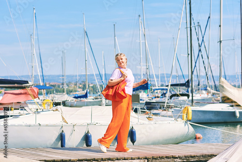 Outdoor fashion portrait of middle age 50 - 55 year old womn wearing colorful outfit, posing at the lake or sea port, travel and vacation lifestyle © annanahabed