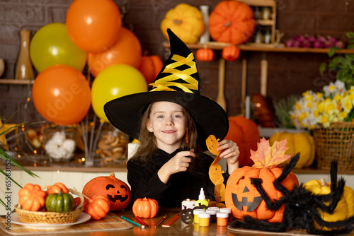 halloween, a child girl in a witch costume with pumpkins and a big spider in a dark kitchen cuts a festive garland with scissors, smiles, rejoices