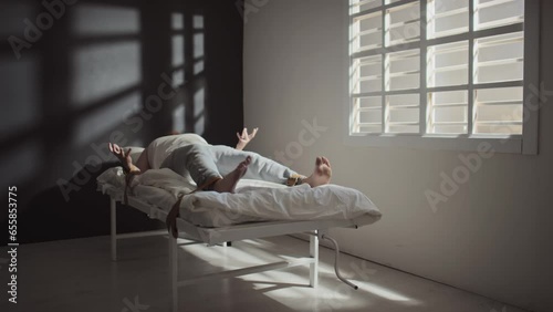 Long shot of Caucasian man with mental disorder lying on hospital bed tied to it with belts, suffering seizure
