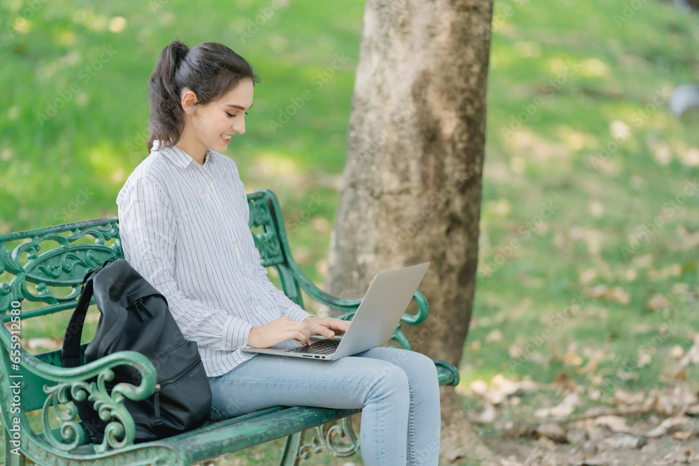 Smiling young woman works online using a laptop in leafy park. She is happily answering chats with customers who are interested in her products. Smiling young woman works online using a laptop in leafy park. She is happily answering chats with customers who are interested in her products.