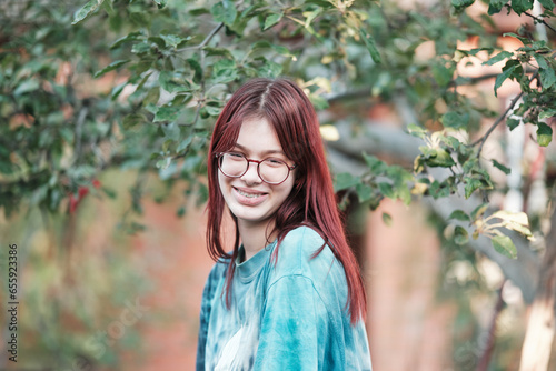 Beautiful red-haired girl with glasses in a turquoise T-shirt smiles against the background of trees in the garden.