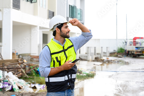 portrait of young engineer in vest with white helmet standing on construction site, smiling and holding smartphone for worker, internet, social media. photo