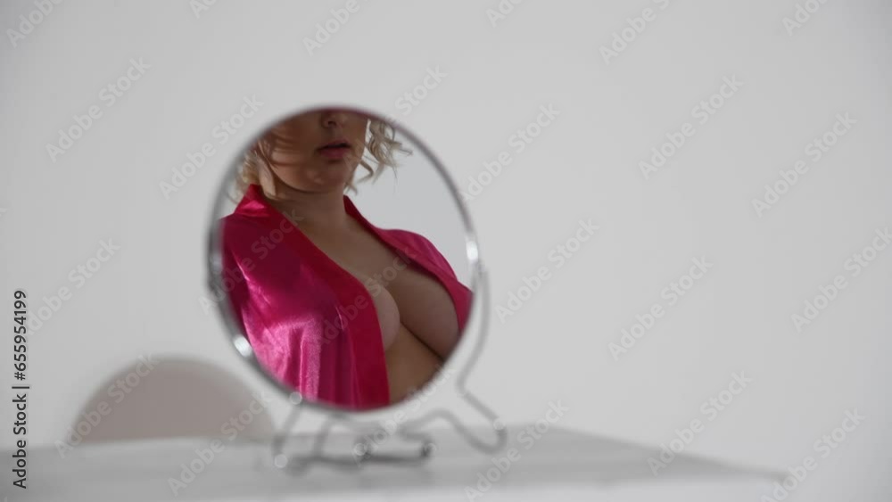 Small round mirror stands on white table in white room. Sexy caucasian plus size woman wearing pink robe on naked body reflects on mirror surface. Real time video. Abstract sensual background.